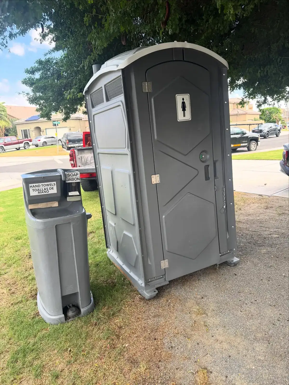 Grey standard porta potty next to a two-sided hand-washing station